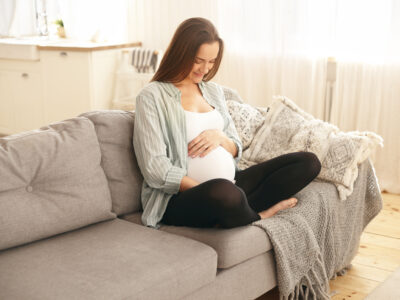 Adorable cute young pregnant woman in casual clothes sitting comfortably on sofa with hands on her belly, talking to her unborn son, smiling happily, enjoying sweet moments of first pregnancy